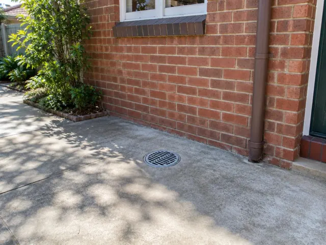 Concrete patio with a round outdoor drain grate beside a brick house wall in Normanhurst